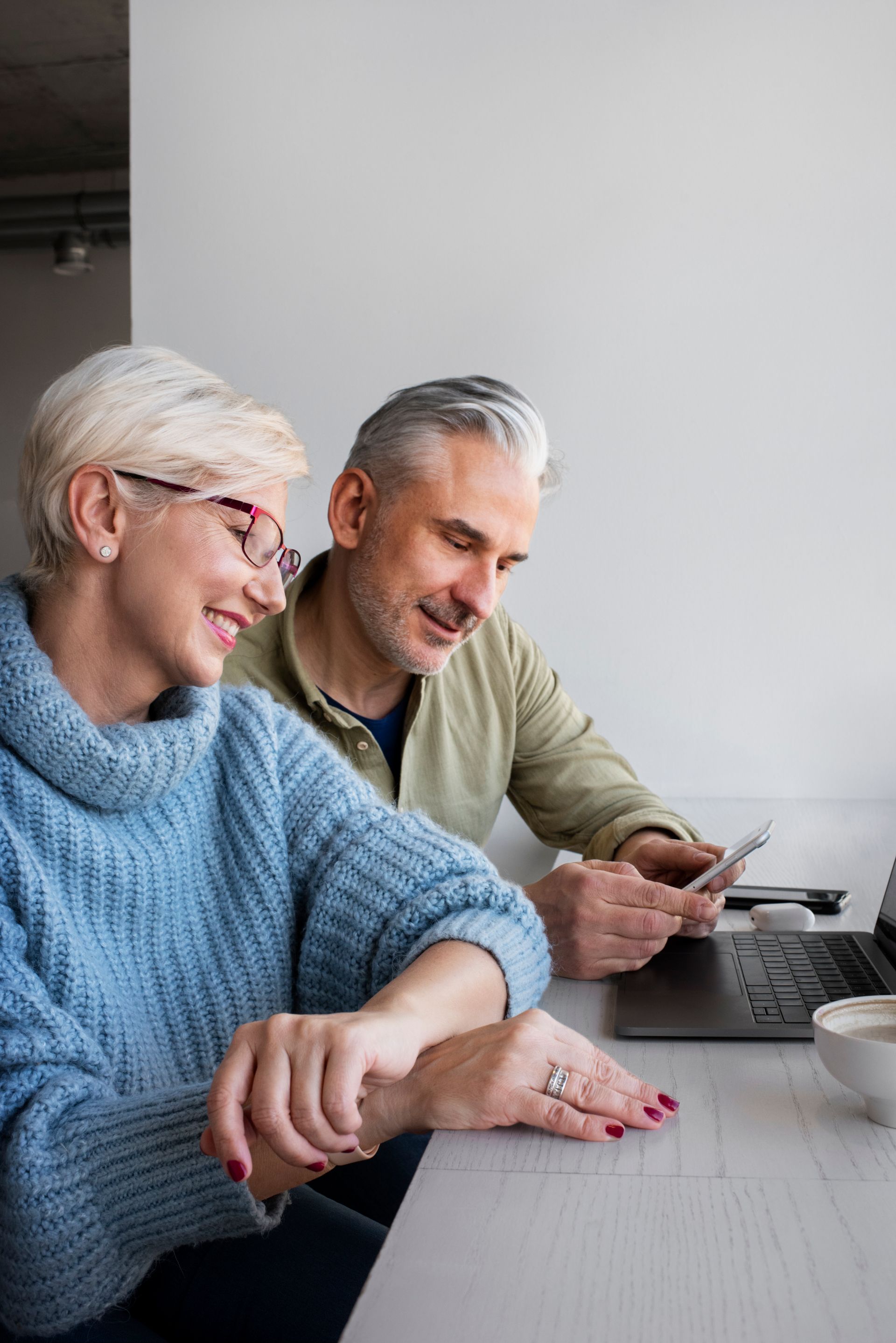 Couple smiles while looking at a phone and laptop on a table. One wears glasses and a blue sweater.