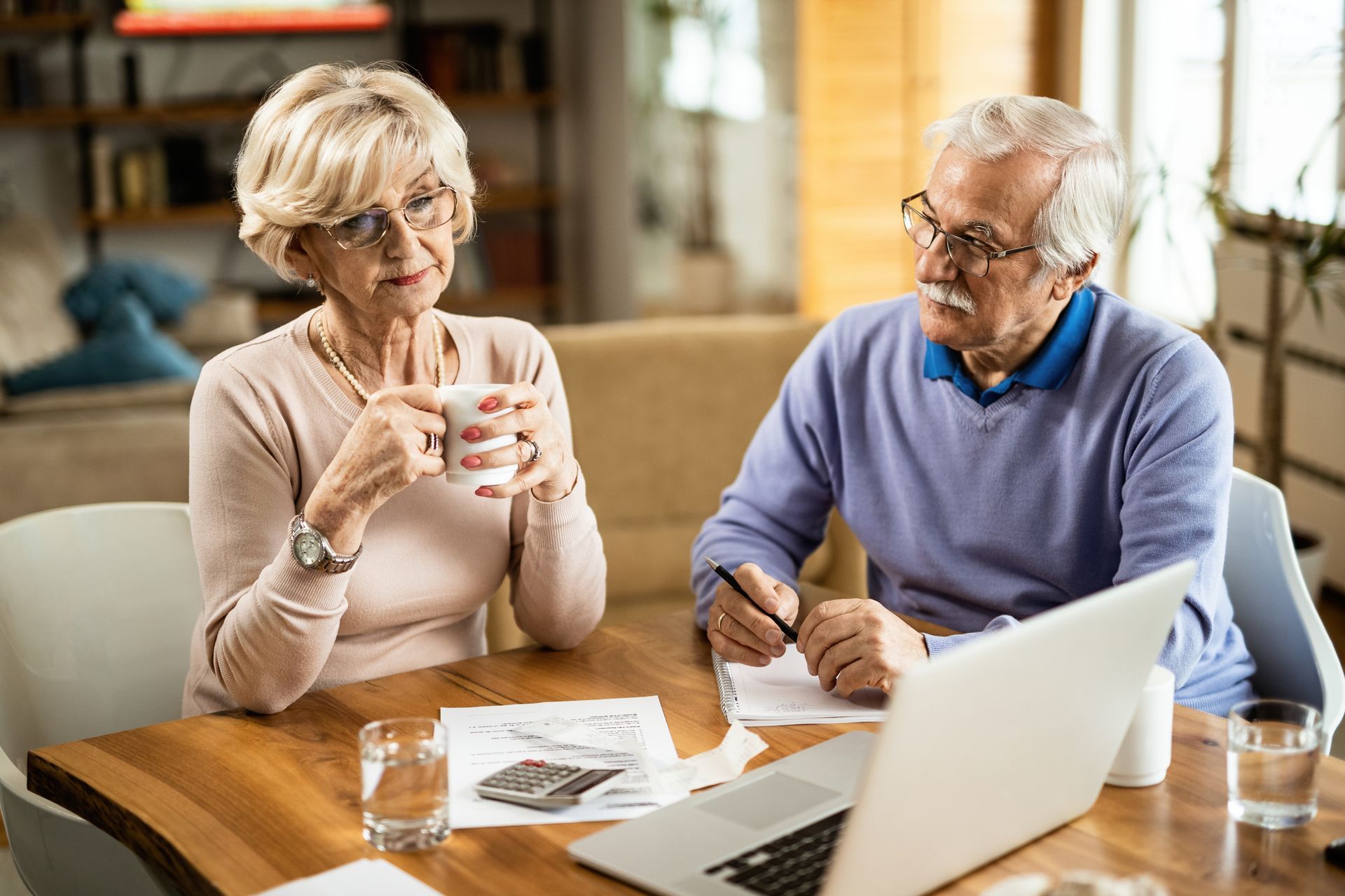 Elderly couple reviews finances at a table with laptop, papers, and coffee.