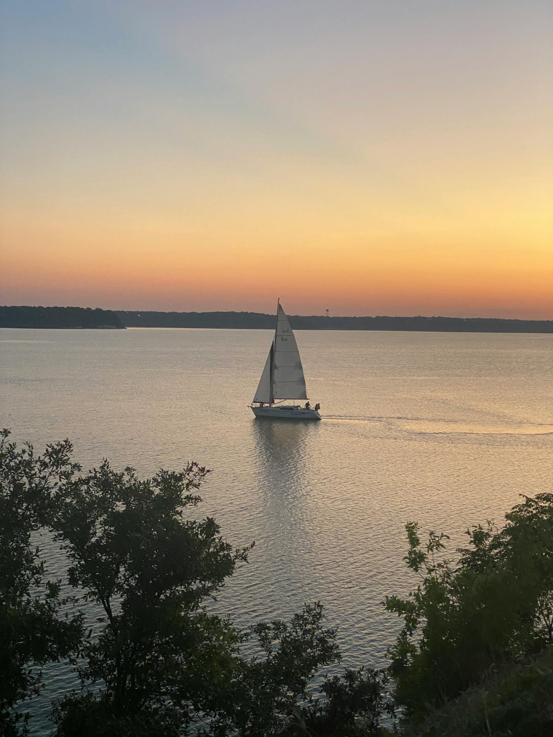 Sailboat on calm water at sunset, orange and blue sky, trees in foreground.