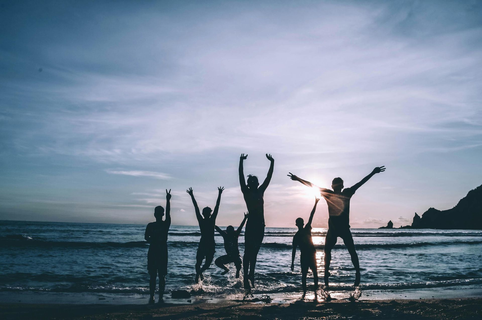 Silhouetted group jumps on a beach at sunset, arms raised. Water and sky in background.