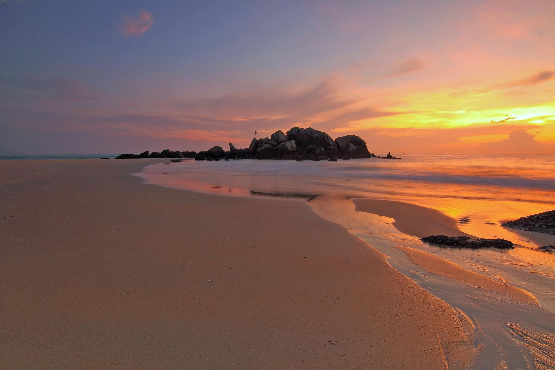 Beach at sunset, water reflecting orange and pink sky. Island silhouette in distance.