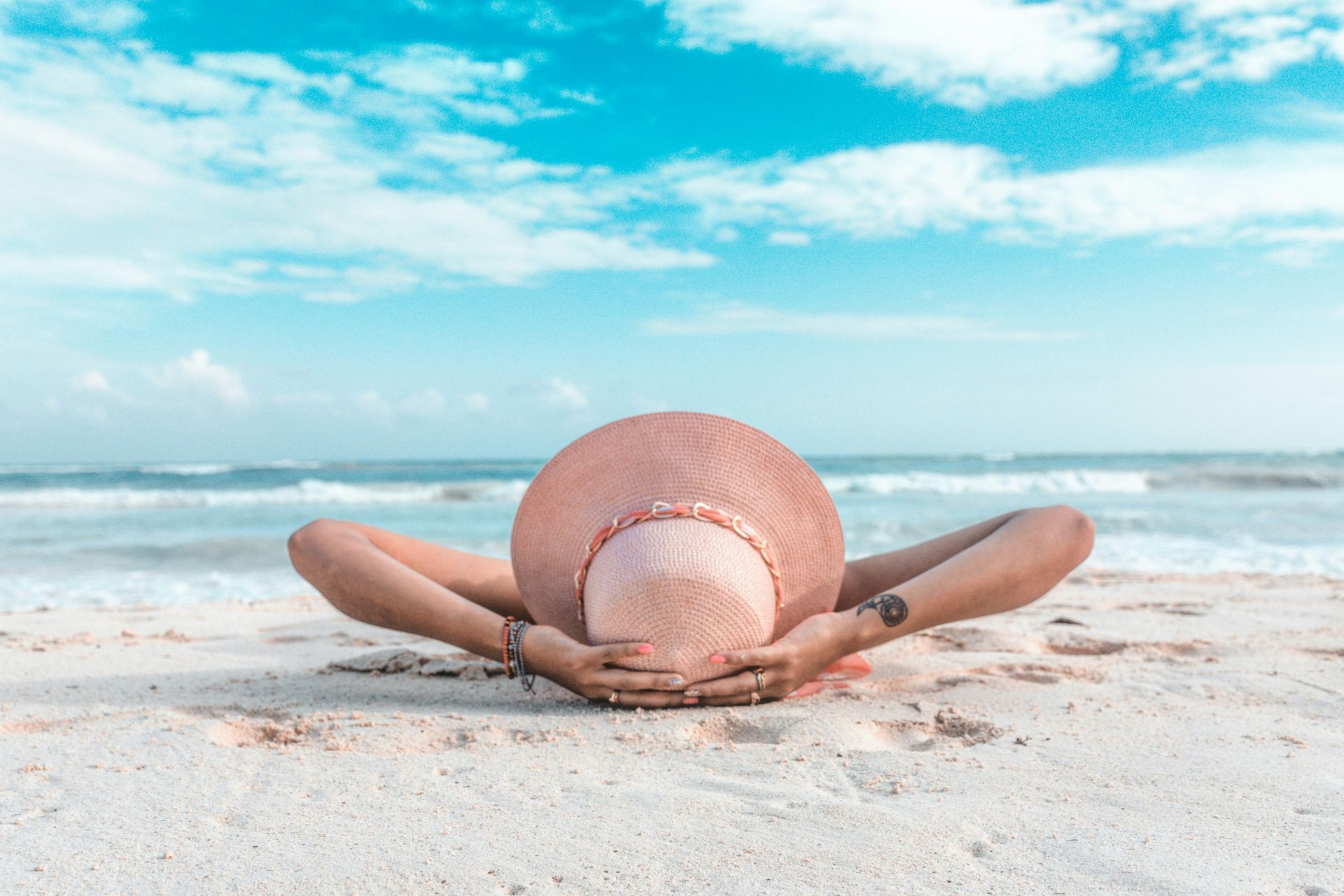 Person wearing a pink hat sunbathing on a white sandy beach with turquoise water.