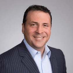 Man in a blue pinstripe suit, smiling, with a light blue shirt, against a gray background.