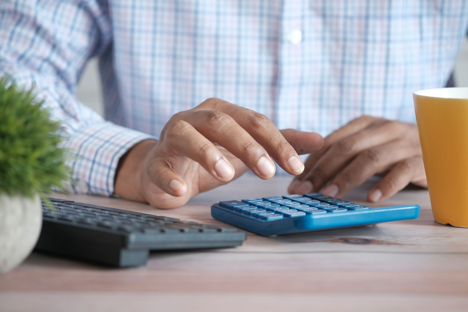 Hands using a blue calculator on a desk with a keyboard, plant, and mug.