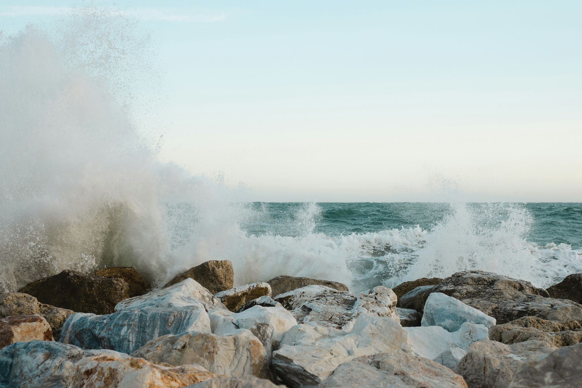 Ocean waves crashing against a rocky shore, creating white spray against a light blue sky and turquoise water.