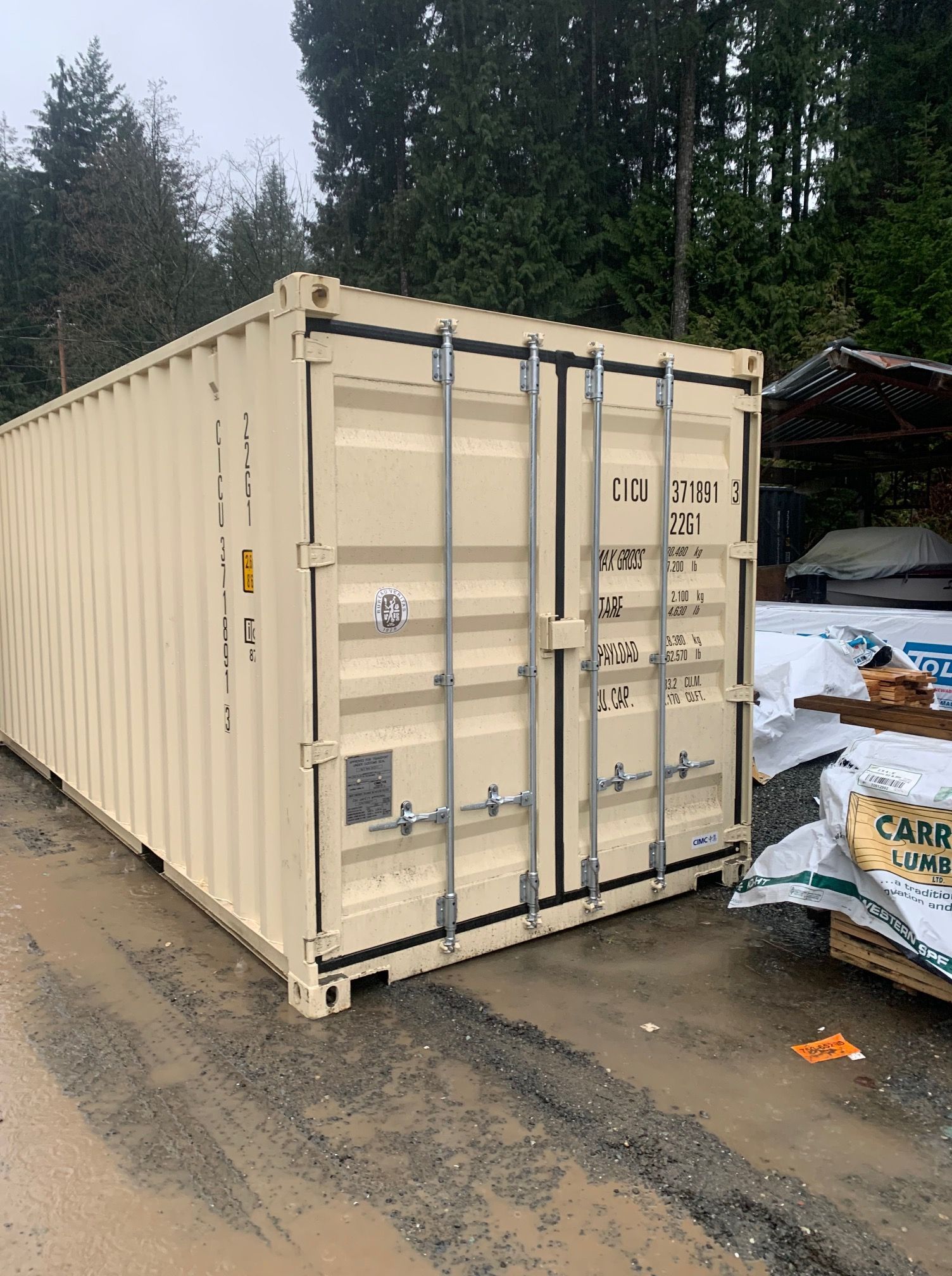 Beige shipping container outdoors on a gravel surface, near trees and lumber.
