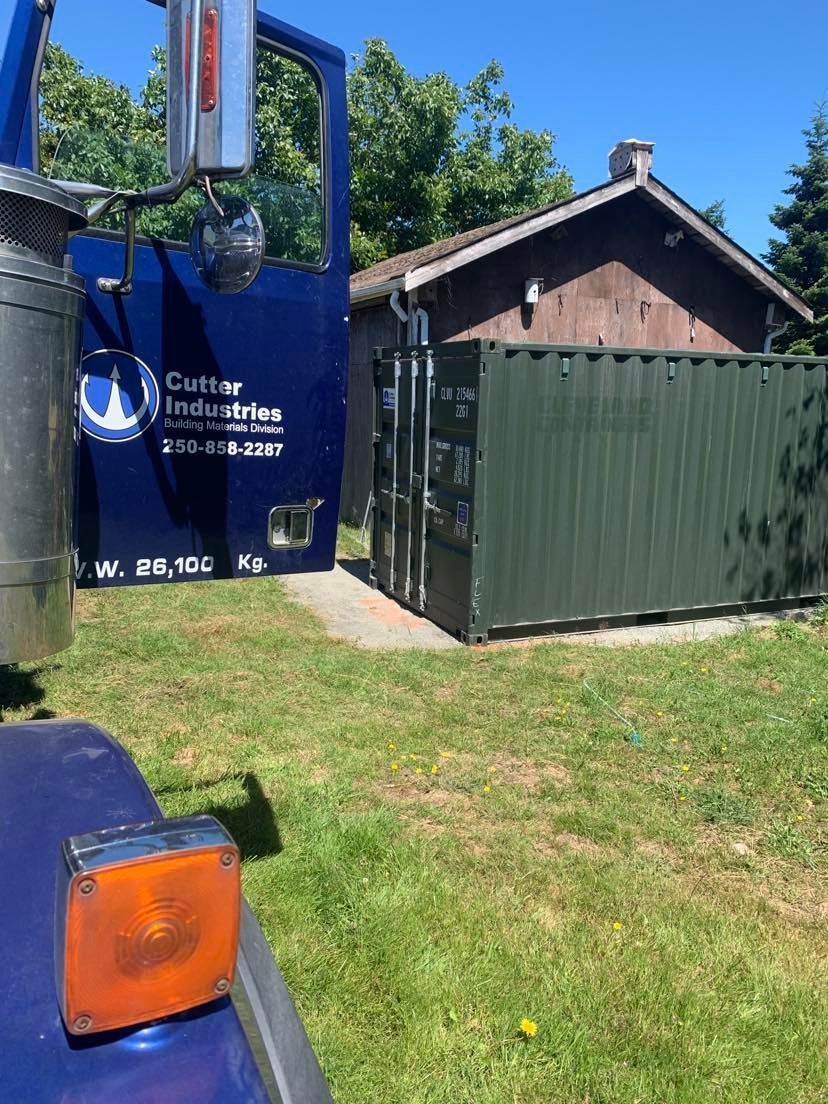 Blue truck next to green shipping container on grass, with a wooden shed in the background.