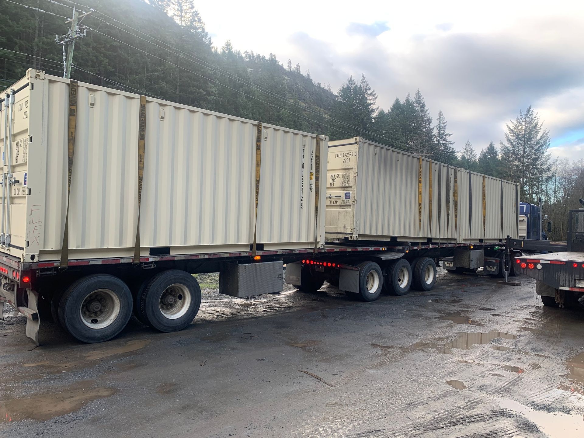 Two beige shipping containers loaded on a semi-truck trailer parked on gravel near trees.