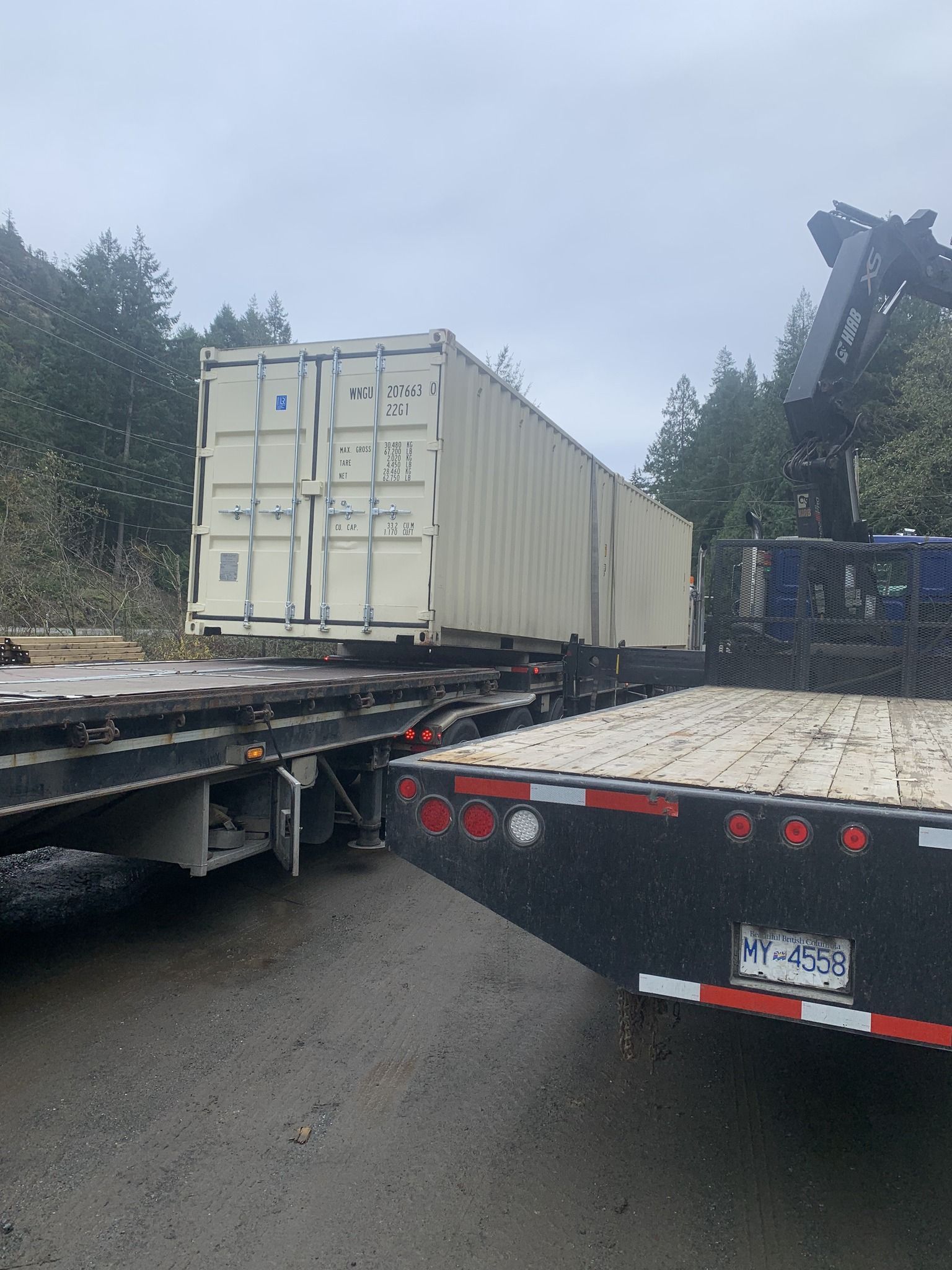 Shipping container on a flatbed trailer, with a crane nearby, in a forest setting.