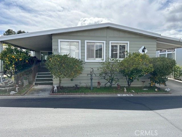 A light green manufactured home with horizontal siding, a covered carport, and small trees in the front yard.