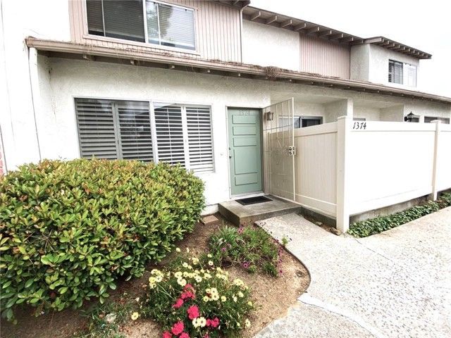 A view of a two-story residential building entrance with a pale green door, a large bush, and a white privacy fence.