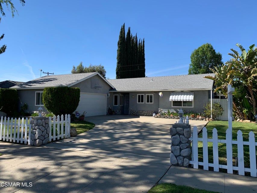 A one-story gray house with a white fence, a stone-pillar gate, and a concrete driveway under a clear blue sky.