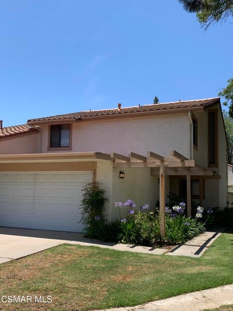 A two-story tan residential building with a white garage, tile roof, and a wooden pergola over the front entryway.