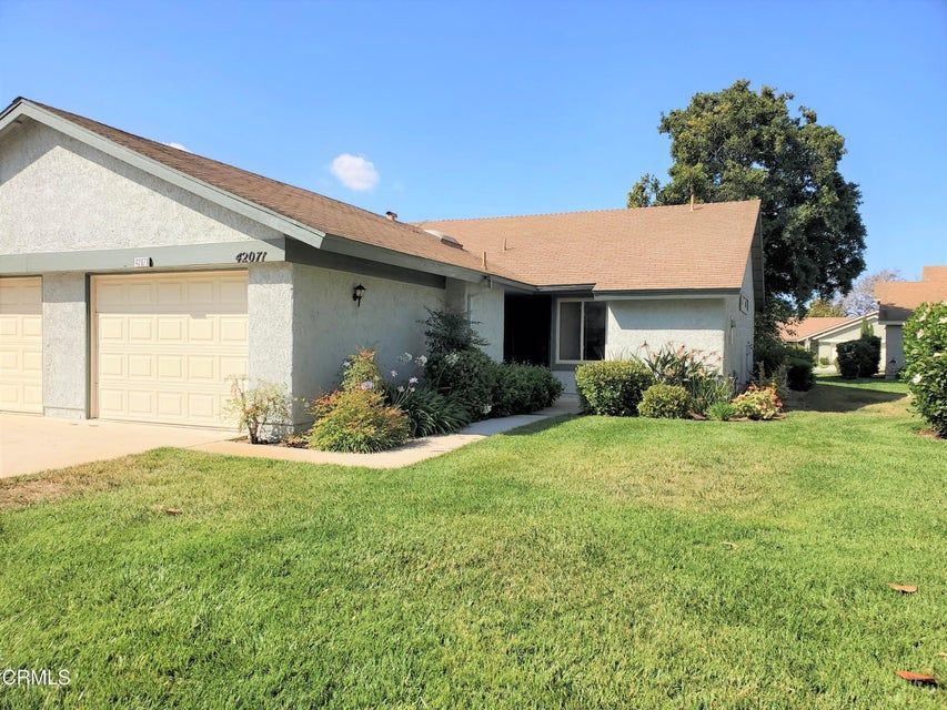 A light-colored single-story suburban house with a tan garage and a neatly mowed front lawn under a clear blue sky.