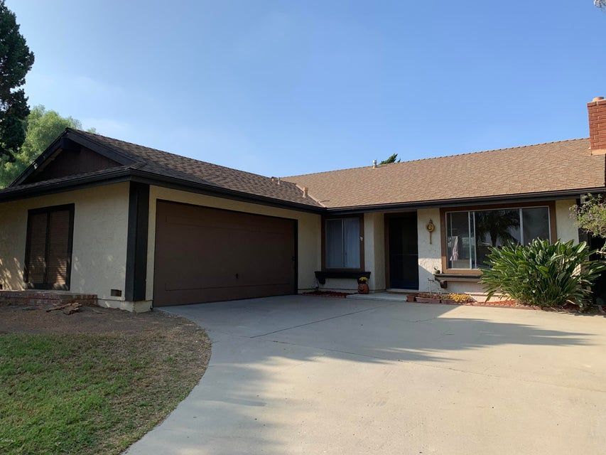 A single-story tan house with a brown roof and matching garage, featuring a paved driveway and a front yard lawn.