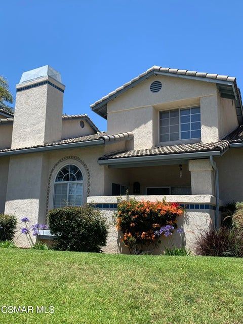 A two-story beige stucco house with a tiled roof, chimney, arched window, and front porch, set against a blue sky.