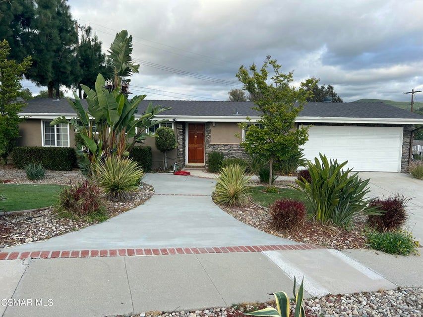 A single-story, beige suburban house with a dark roof and white garage, viewed from the street across a concrete driveway.