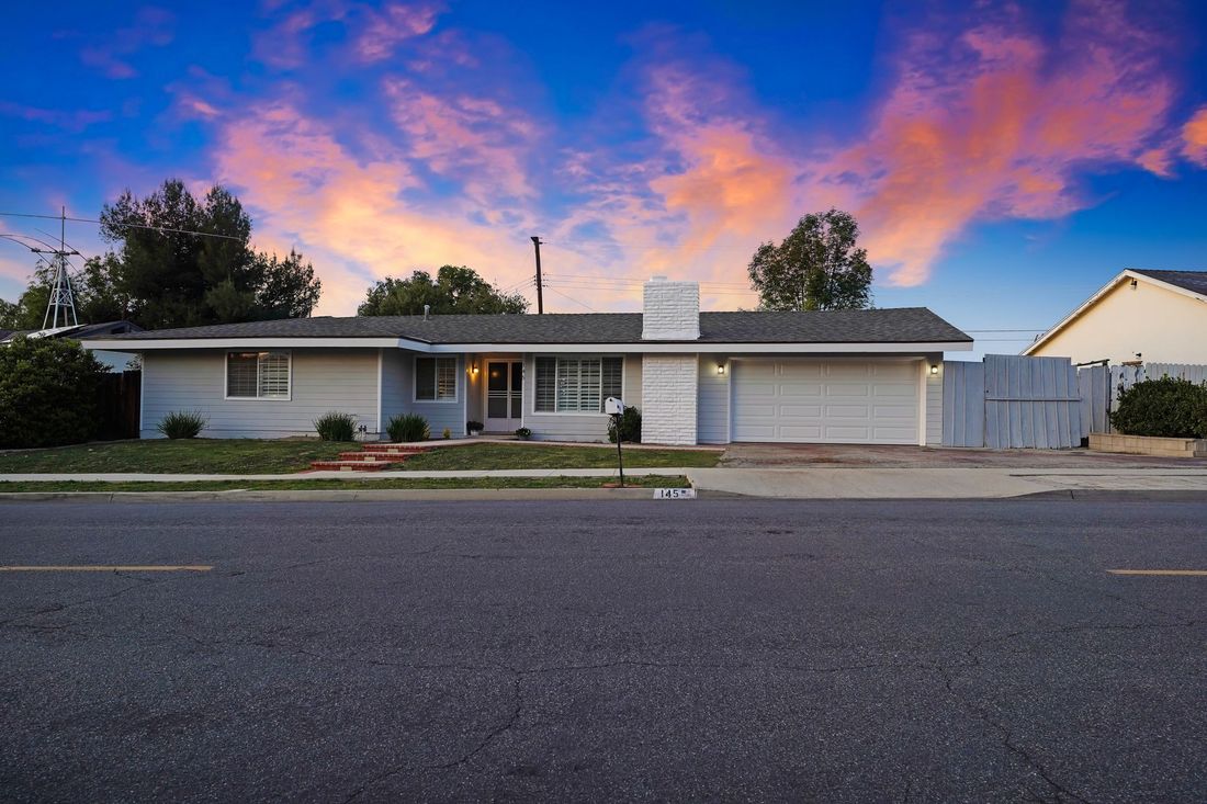 A one-story suburban house with a white garage and brick chimney under a vibrant purple and orange sunset sky.