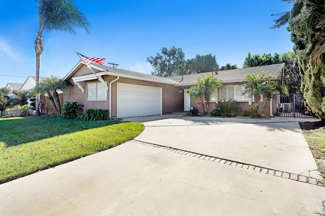A single-story, tan-colored suburban home with a white garage, a concrete driveway, and a small front lawn under blue sky.