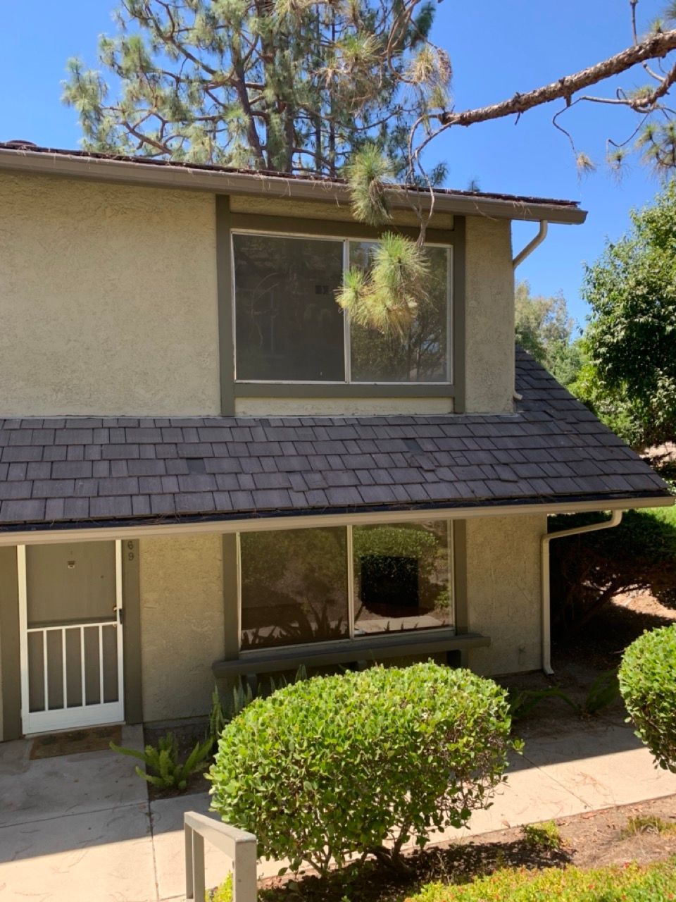 A two-story tan apartment unit with brown shingle siding, a white front screen door, and green manicured shrubs.