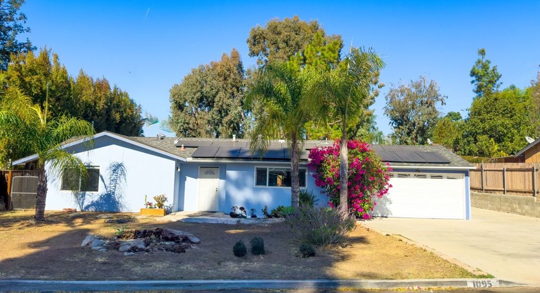 A single-story, light blue suburban house with a white garage door, palm trees, and a desert-style front yard.