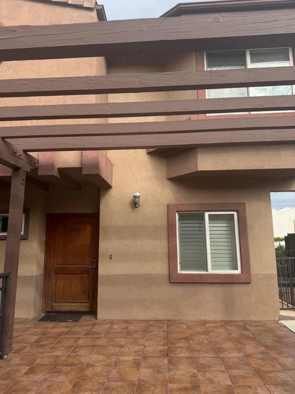 A tan two-story stucco building with a wooden pergola, a dark wood front door, and a tiled patio.