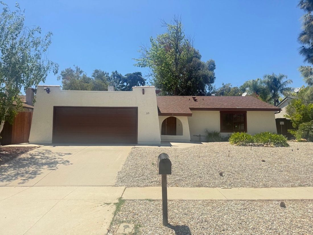 A single-story, tan, Southwestern-style house with a brown roof and garage, featuring a gravel front yard under a blue sky.