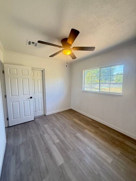 A bright, empty room with gray wood-look flooring and white walls; a closed white door, a window, and a ceiling fan.