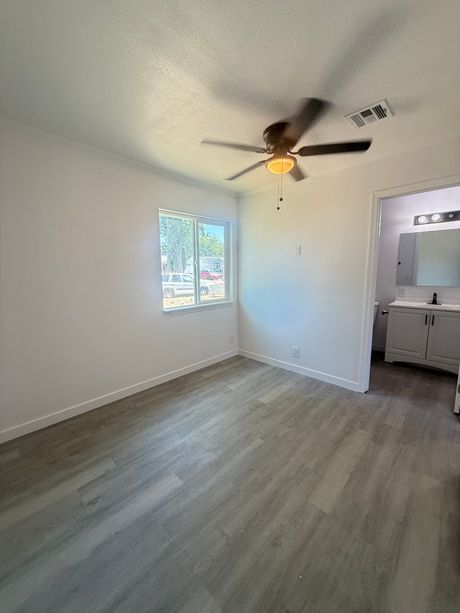 Bedroom with gray wood-look flooring, white walls, window, ceiling fan, and bathroom entrance.