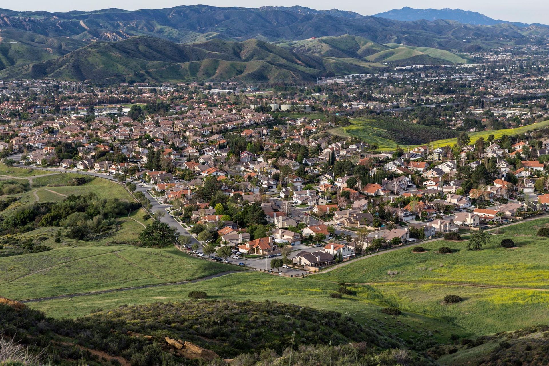 Panoramic view of a suburban town with red-roofed houses, green hills, and mountains in the distance