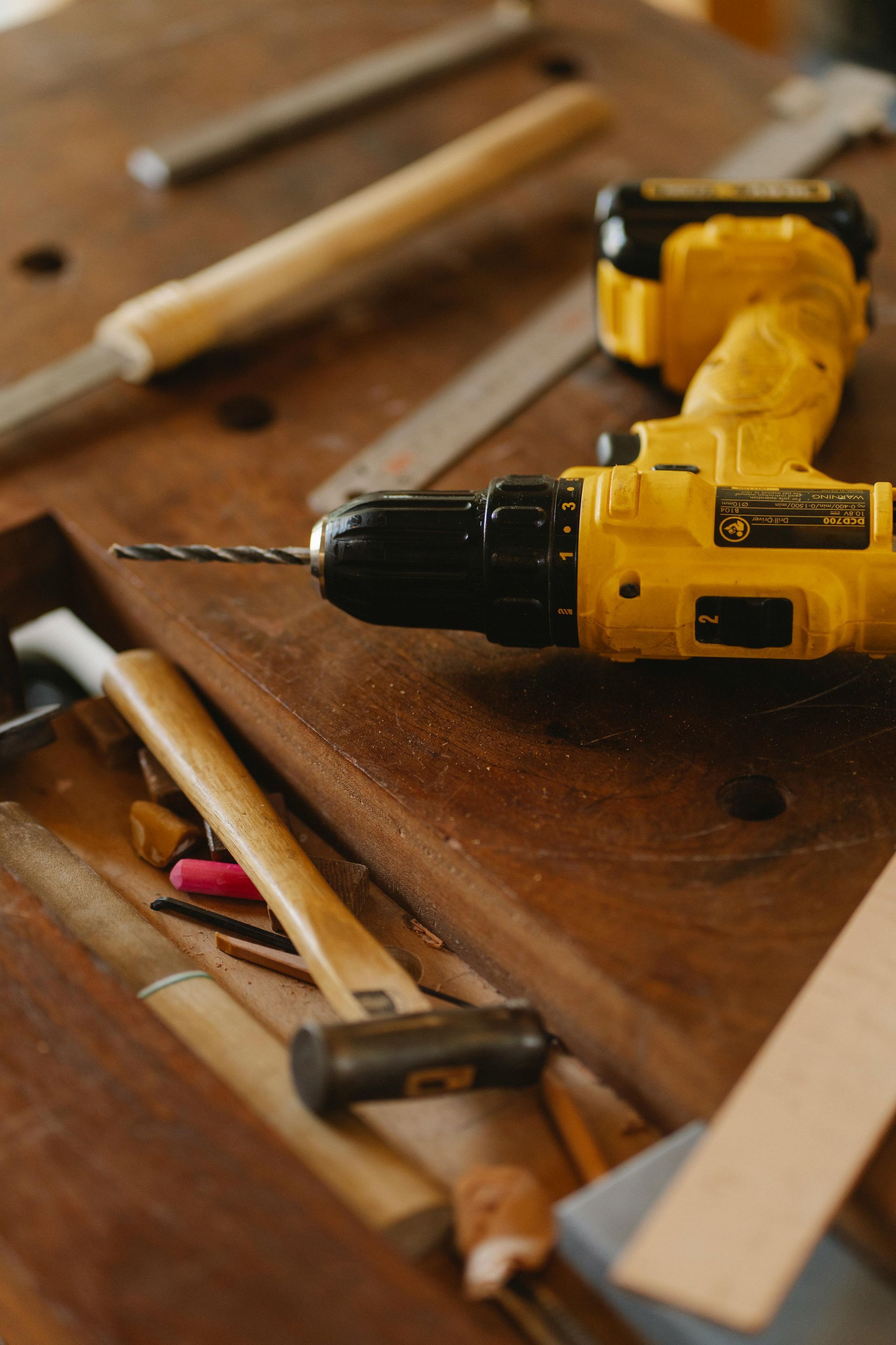 A yellow cordless power drill rests on a wooden workbench surrounded by various workshop tools.