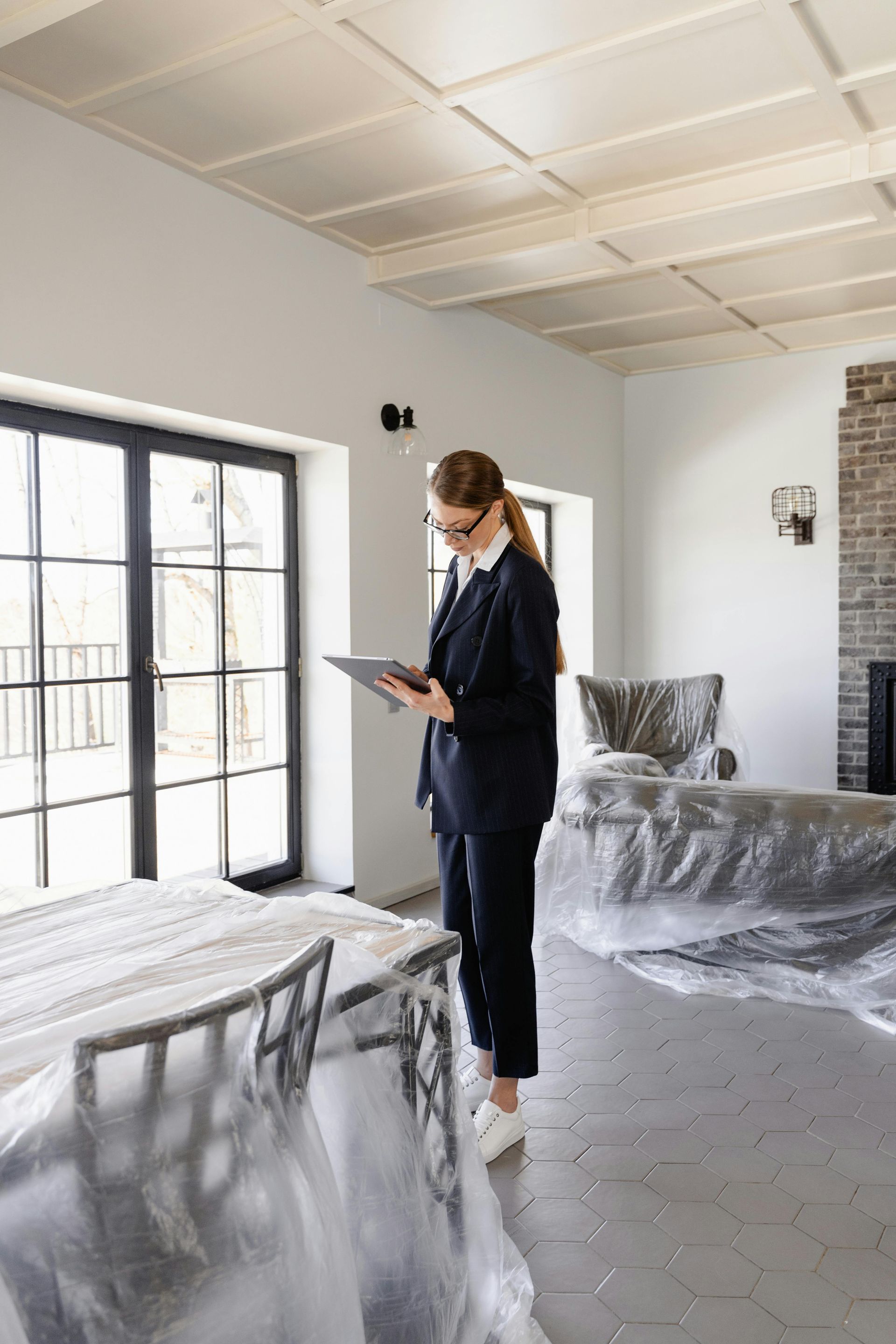 A person in a dark suit uses a tablet while standing in a room with plastic-covered furniture and tiled floors.