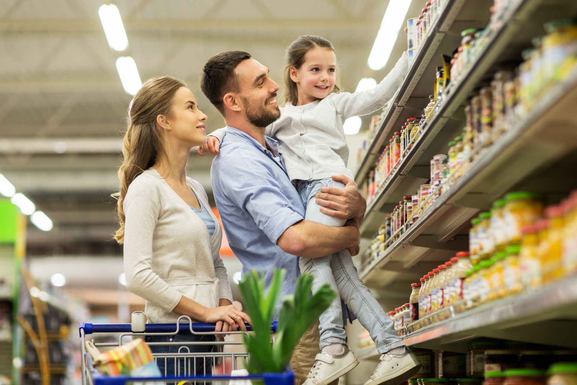 Man in a blue shirt smiling at his phone while grocery shopping with his wife and daughter.