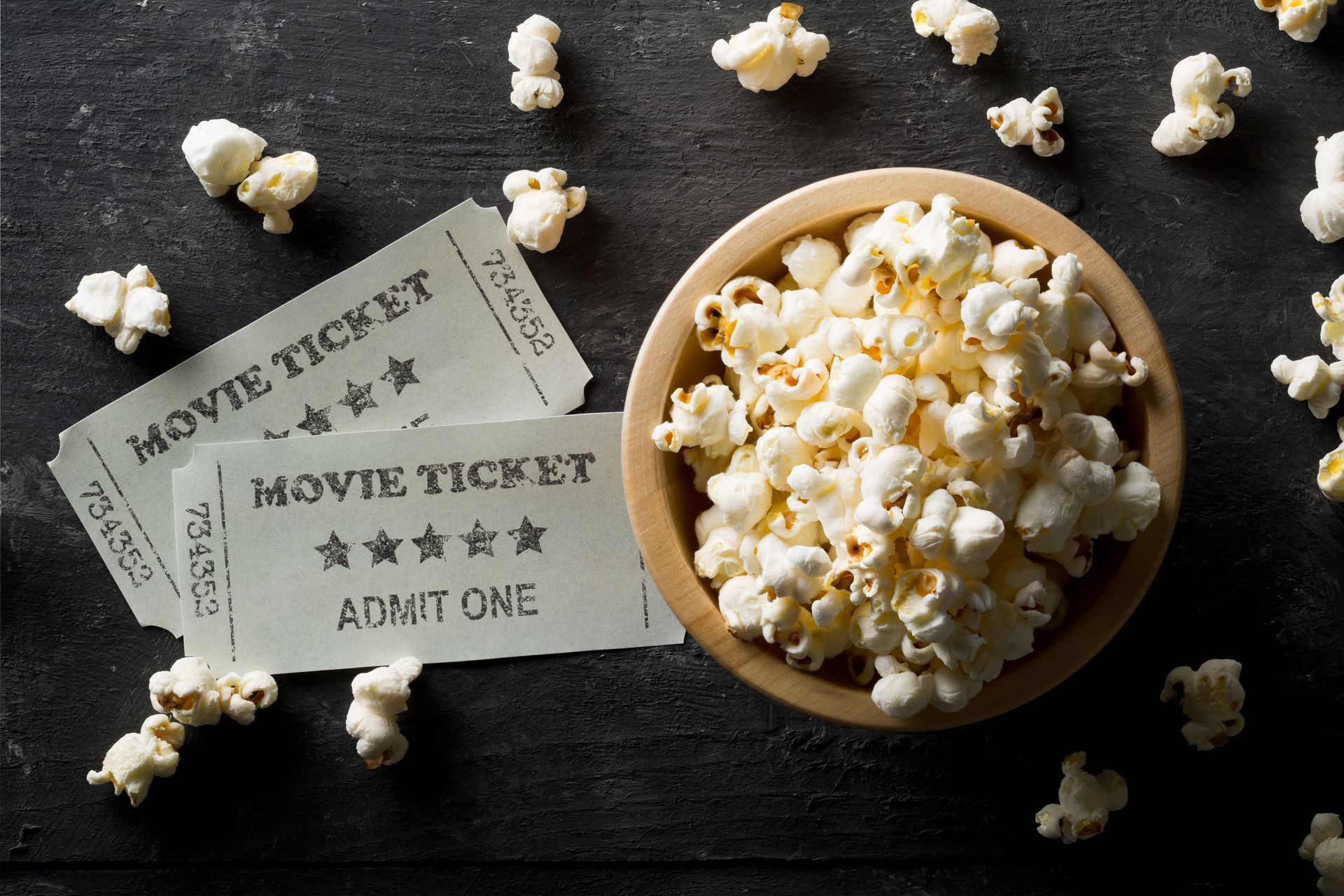 A bowl of popcorn and two movie tickets on a dark surface. Popcorn pieces are scattered around, evoking a playful, cinematic atmosphere.