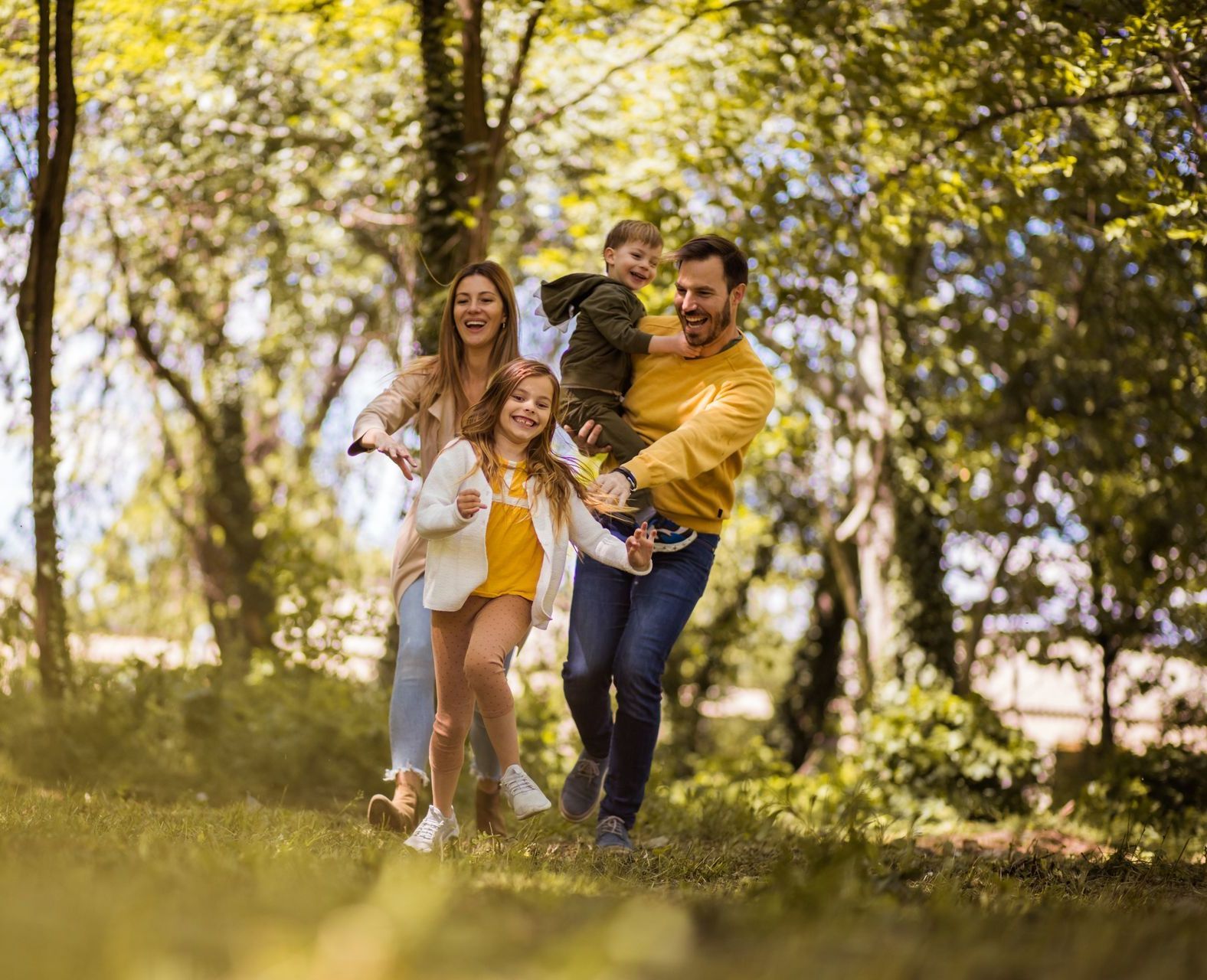 Family of four in a park, smiling, running and being carried among trees, sunshine.