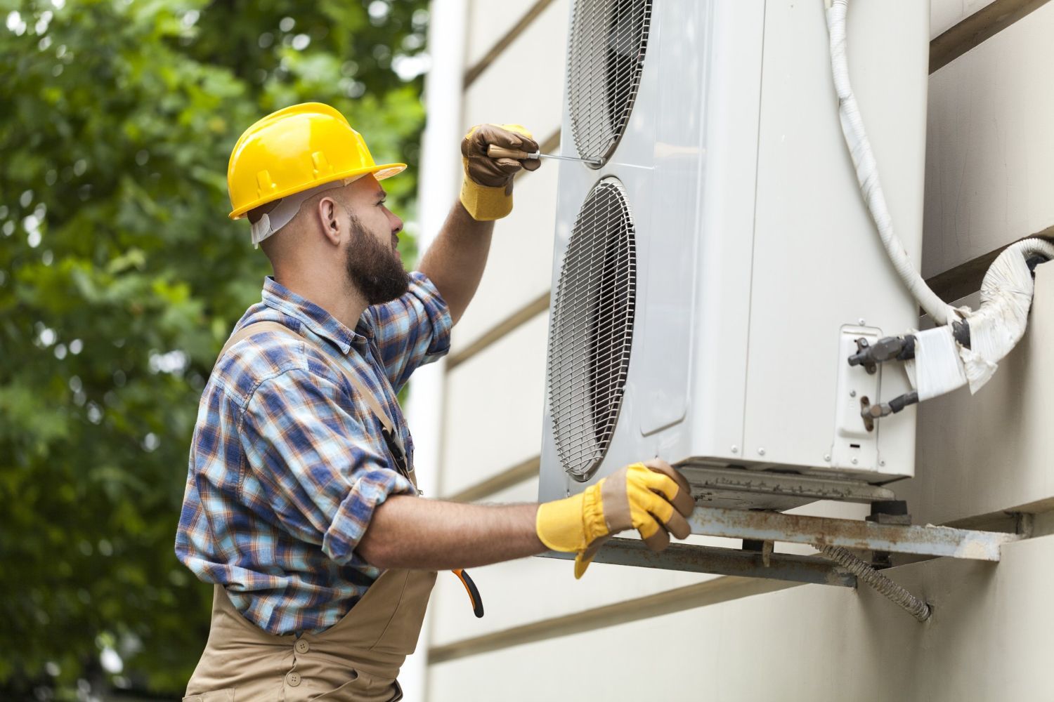 A technician in a yellow hard hat and gloves installs an air conditioning unit on an exterior wall.