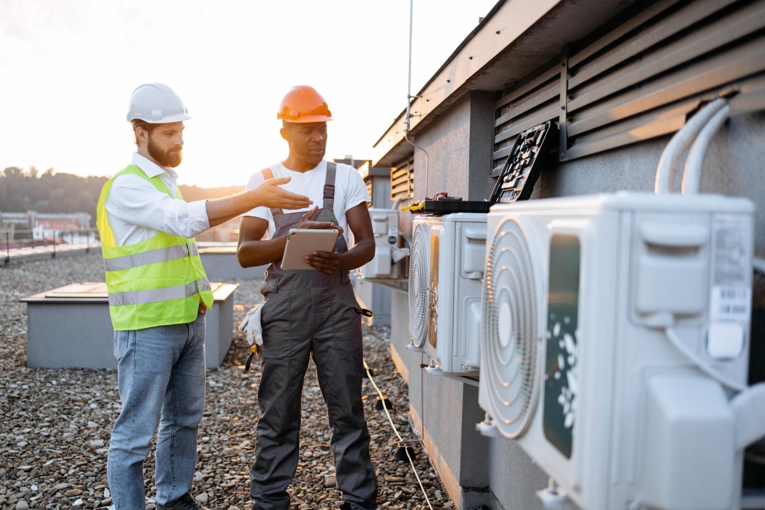 Two workers in safety gear and hard hats discuss HVAC units on a building rooftop at sunset.