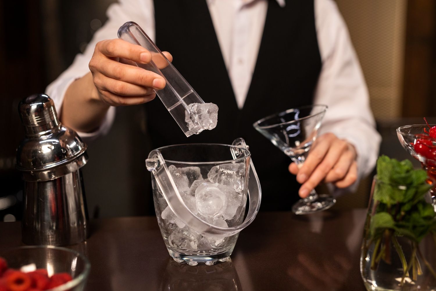 A bartender in a vest uses tongs to place an ice cube into a bucket on a bar counter with a cocktail glass nearby.