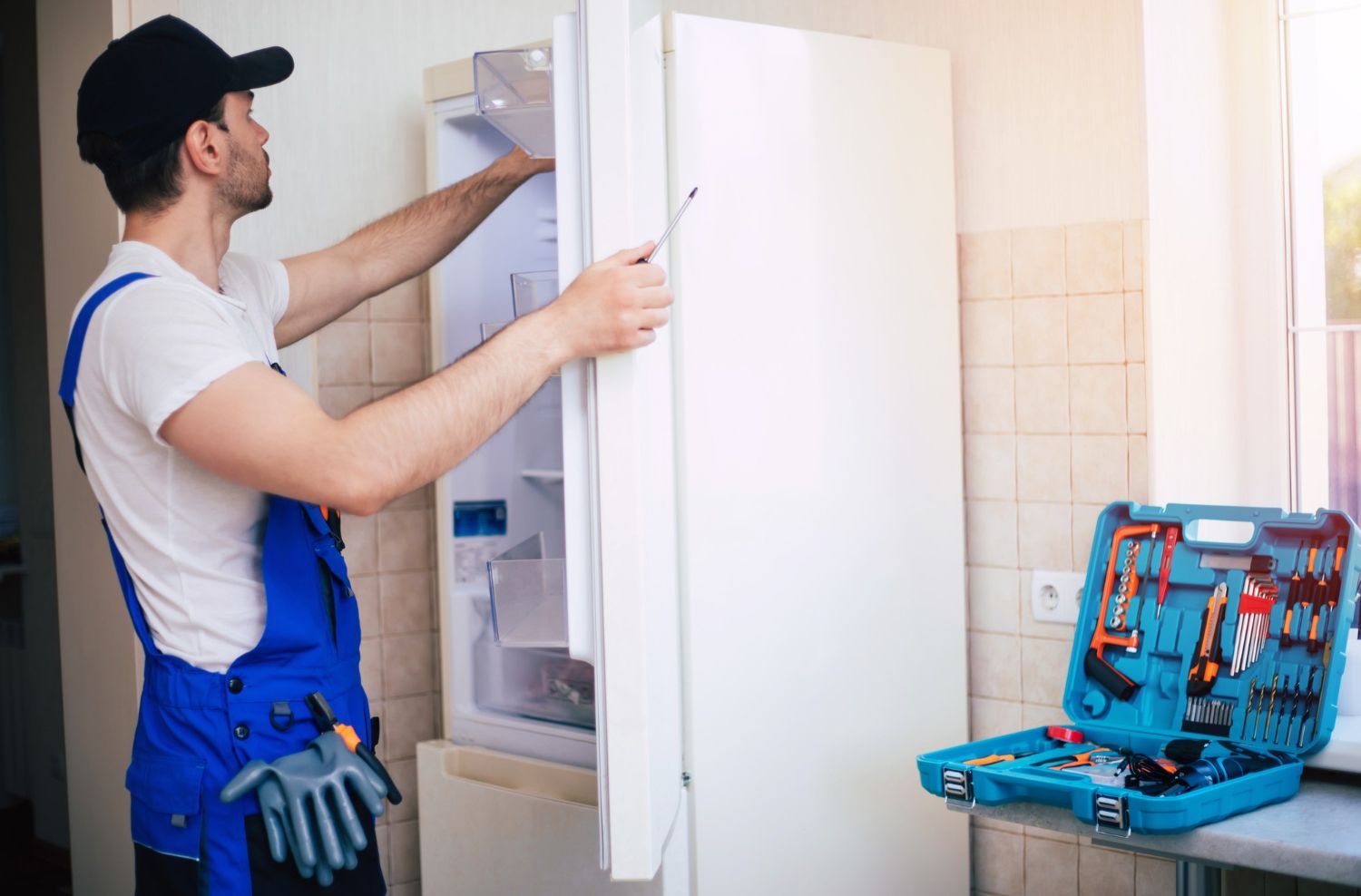 A repair worker in a blue uniform and cap uses a tool to fix a white refrigerator with an open tool kit nearby.