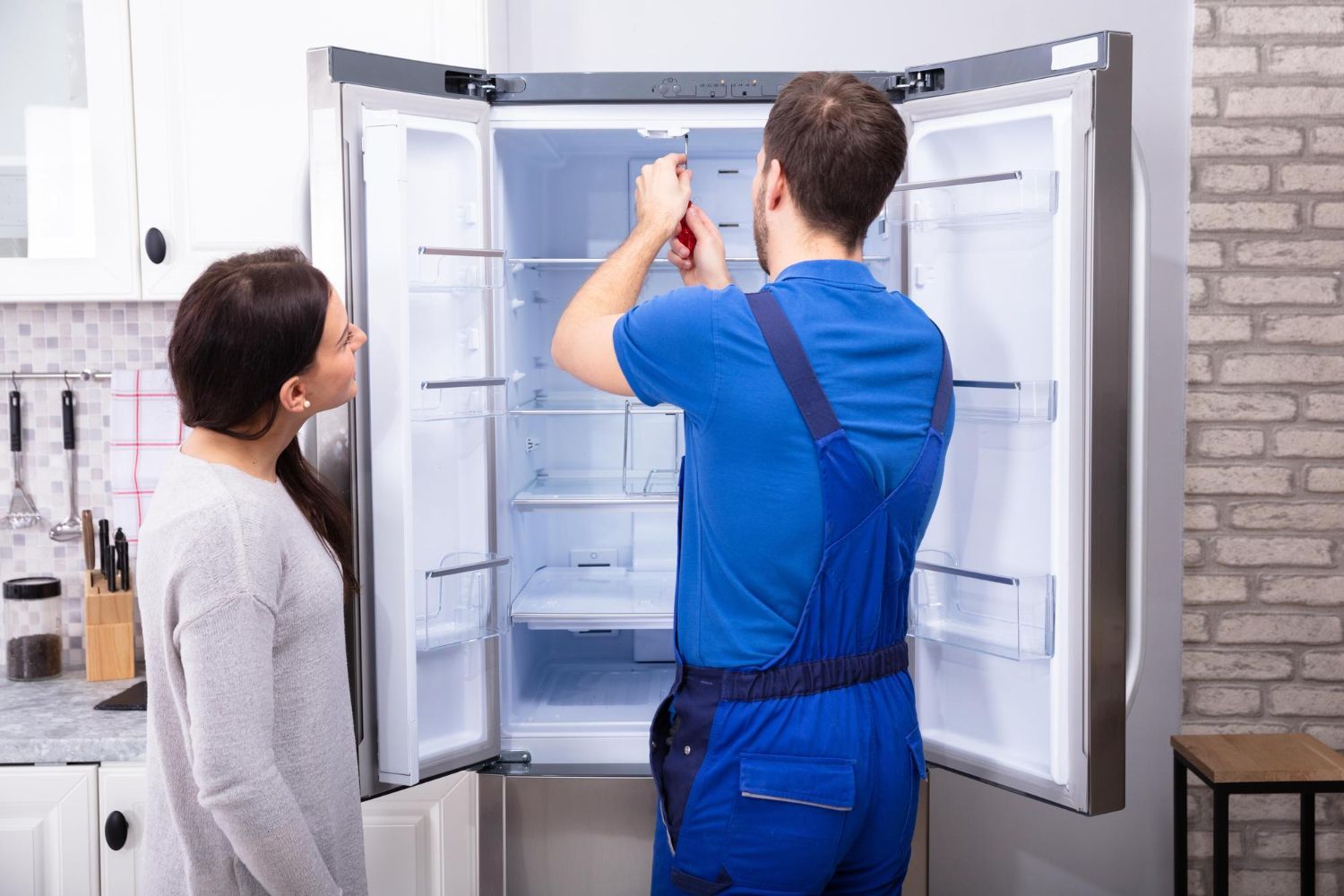 A technician in blue workwear repairs an open stainless-steel refrigerator while a person looks on in a kitchen.