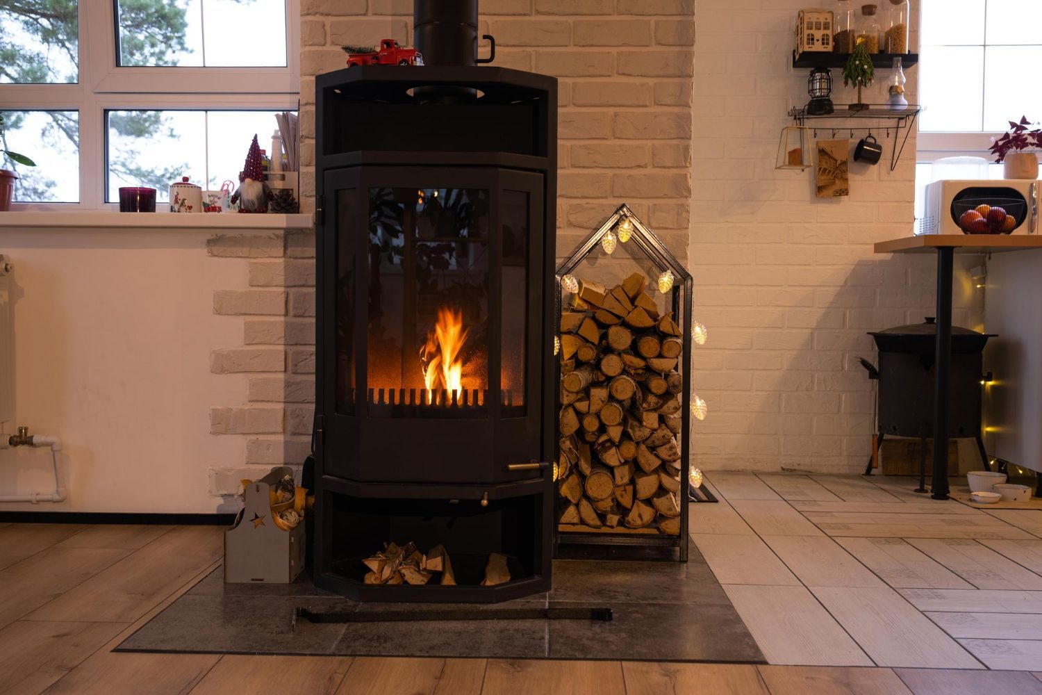 A lit wood stove stands on a tiled hearth next to a house-shaped log holder in a white-walled room with wood flooring.