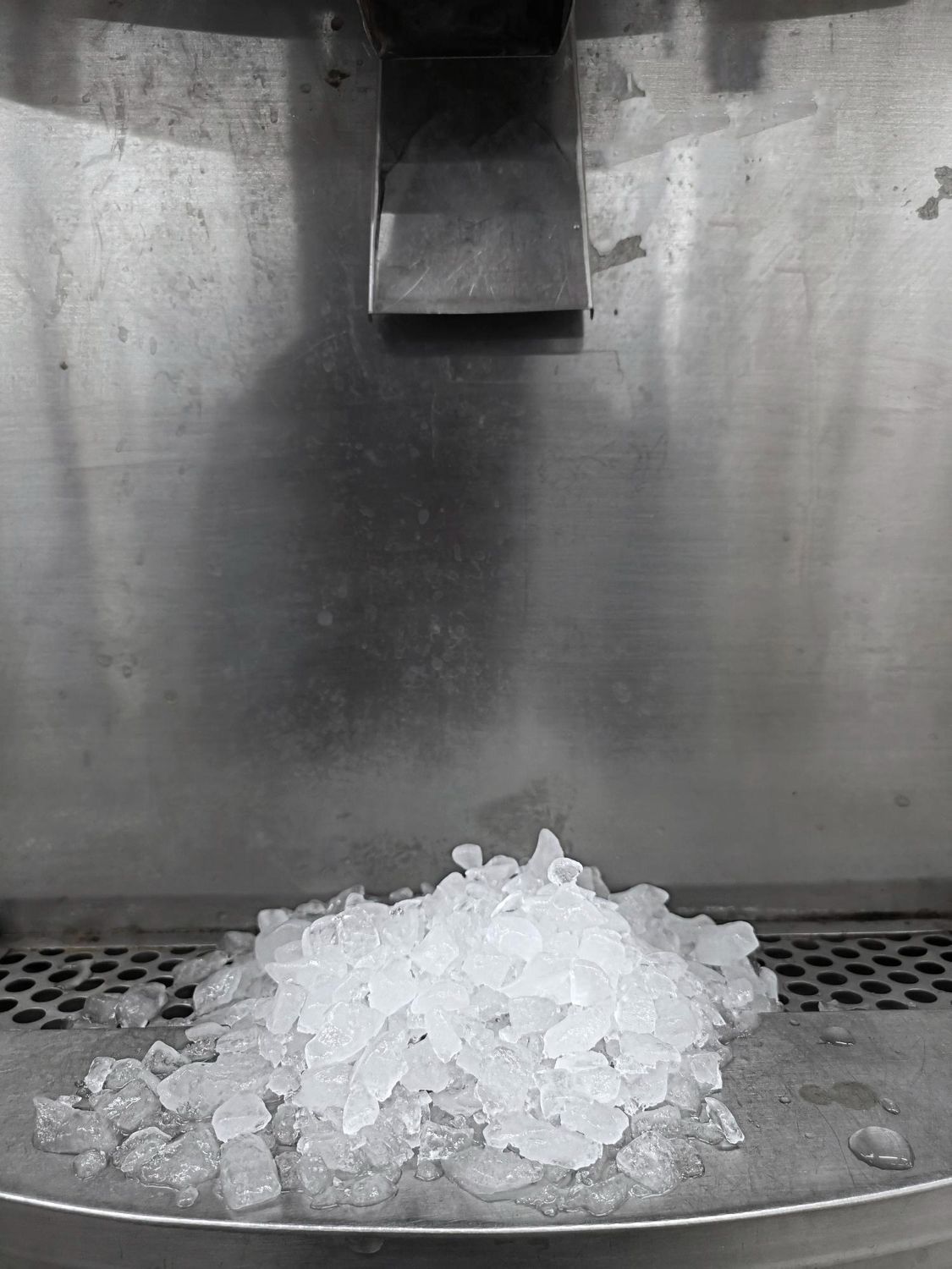A pile of crushed ice sits on the metal drip tray of an ice dispenser.