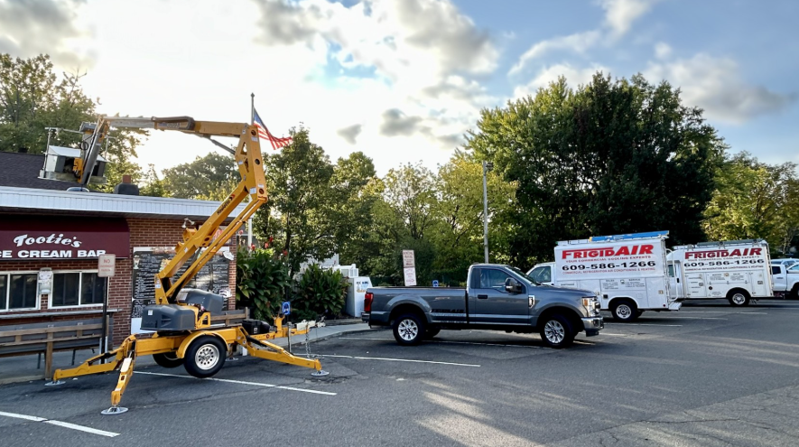 A yellow aerial lift parked next to a storefront, with a pickup truck and two service vans parked in a lot nearby.