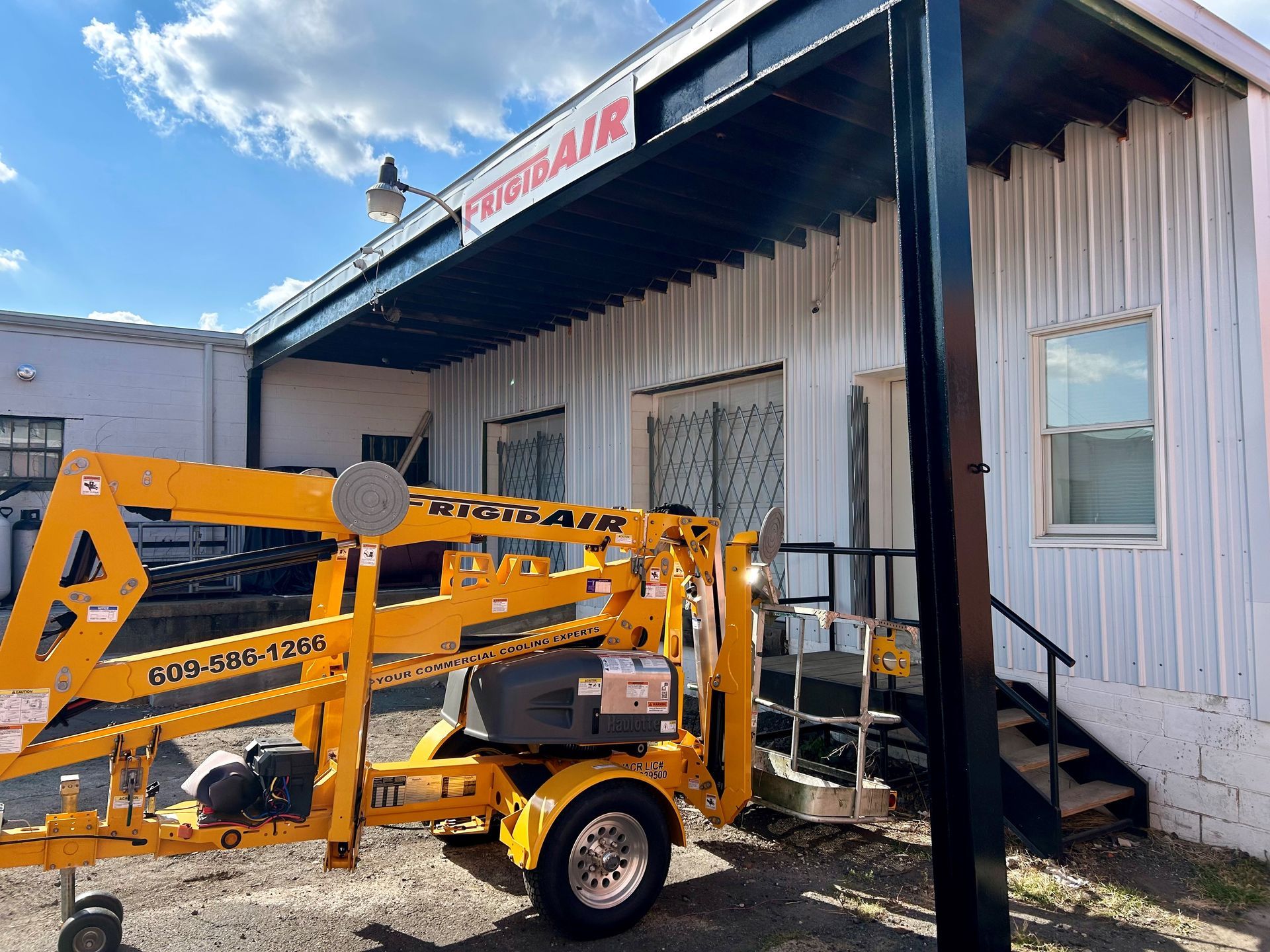 A bright yellow towable boom lift parked in front of a white building with a black awning marked 