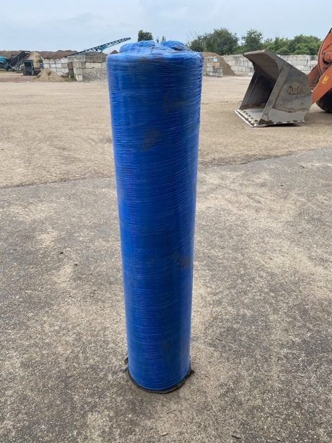 A tall, blue, cylindrical object wrapped in plastic standing upright on a paved surface in an industrial yard.