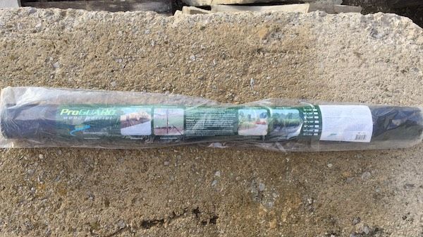 A roll of green plastic garden fencing encased in clear plastic packaging resting on a rough concrete surface.