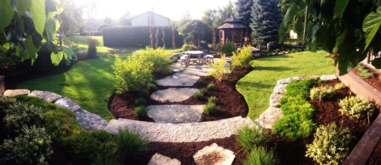 A wide-angle backyard garden with a stone path leading through mulch beds to a gazebo, surrounded by green lawns and shrubs.