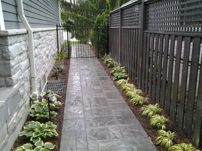 A concrete walkway leads between a gray stone house wall and a wooden privacy fence to a metal gate.