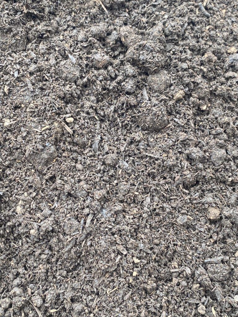 A close-up view of dark, textured soil with organic matter, small clumps, and wood fragments scattered throughout.