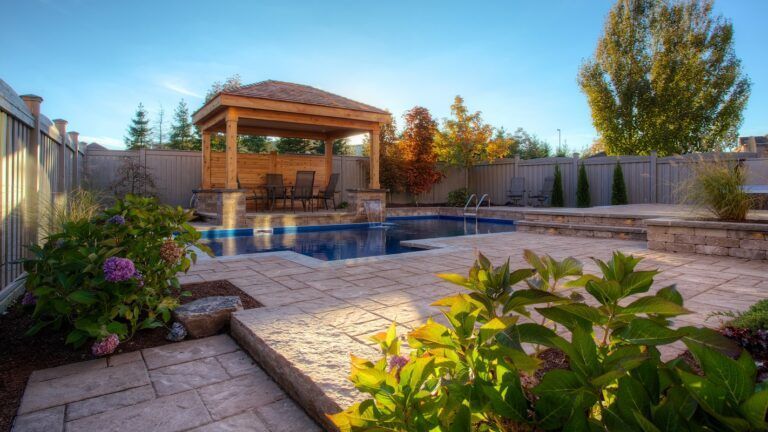 A sunny backyard featuring a rectangular swimming pool with a wooden gazebo and patio area, surrounded by lush landscaping.